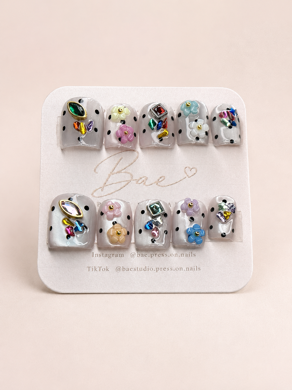 A set of 10 press-on nails displayed on a white card labeled 'Bae'. The nails feature a white base with black polka dots and various colorful rhinestone and floral embellishments.