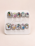 A set of 10 press-on nails displayed on a white card labeled 'Bae'. The nails feature a white base with black polka dots and various colorful rhinestone and floral embellishments.