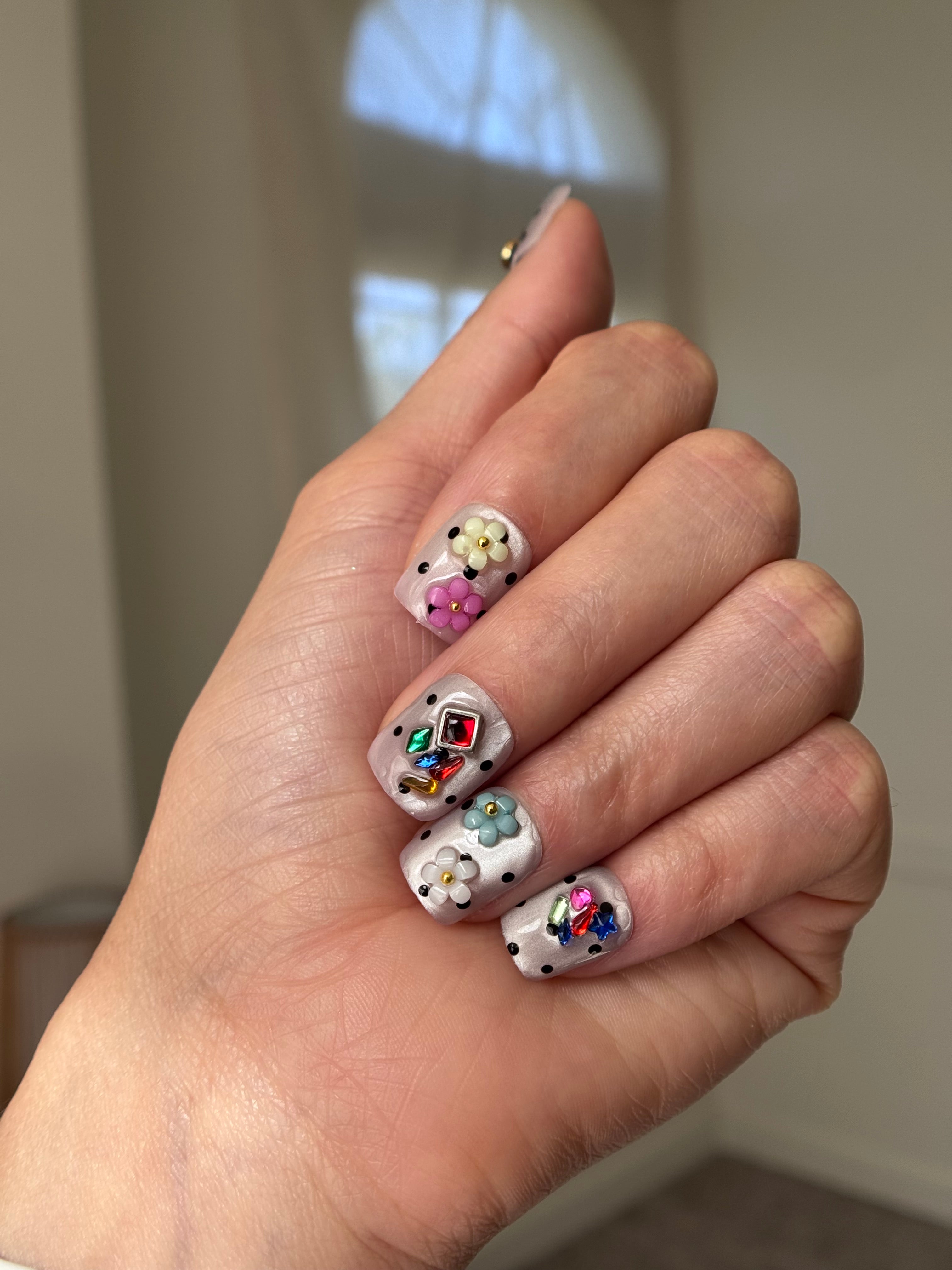A close-up of a hand showcasing intricate nail art designs. The nails feature a white base decorated with small white flowers, colorful rhinestones, and black polka dots, set against a softly blurred indoor background.